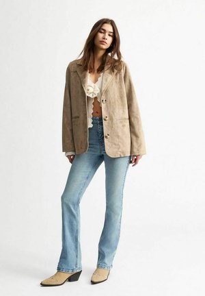 Beige suède blazer met een ontspannen pasvorm, grote kraag en knoopsluiting, gecombineerd met lichtblauwe flared jeans en beige hakken.